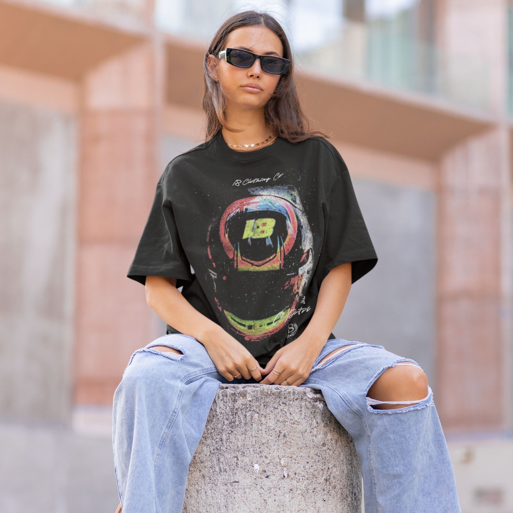Person wearing a black t-shirt with a colorful graphic and ripped jeans, sitting on a concrete block.