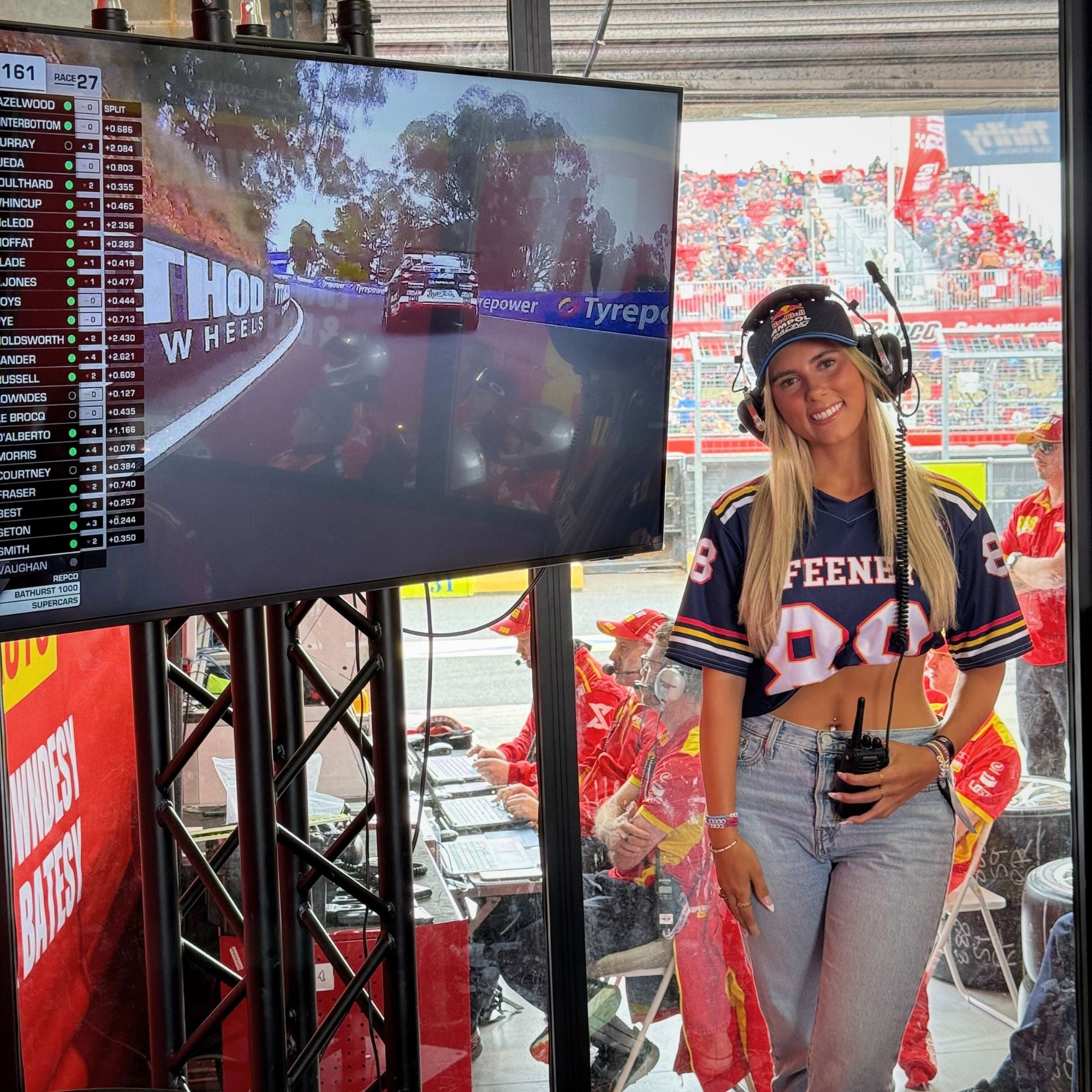 Woman in a sports jersey standing in front of a large screen displaying a race, with a racing event in the background.