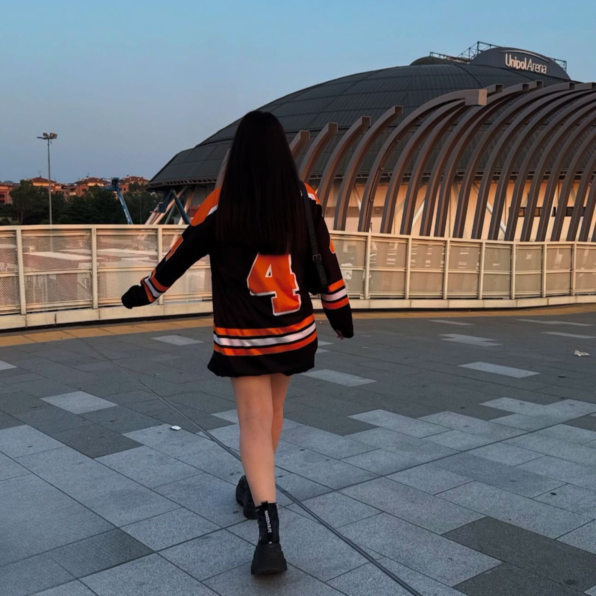 Person wearing a black and orange sweater with number 4, walking on a tiled pavement with a building in the background.