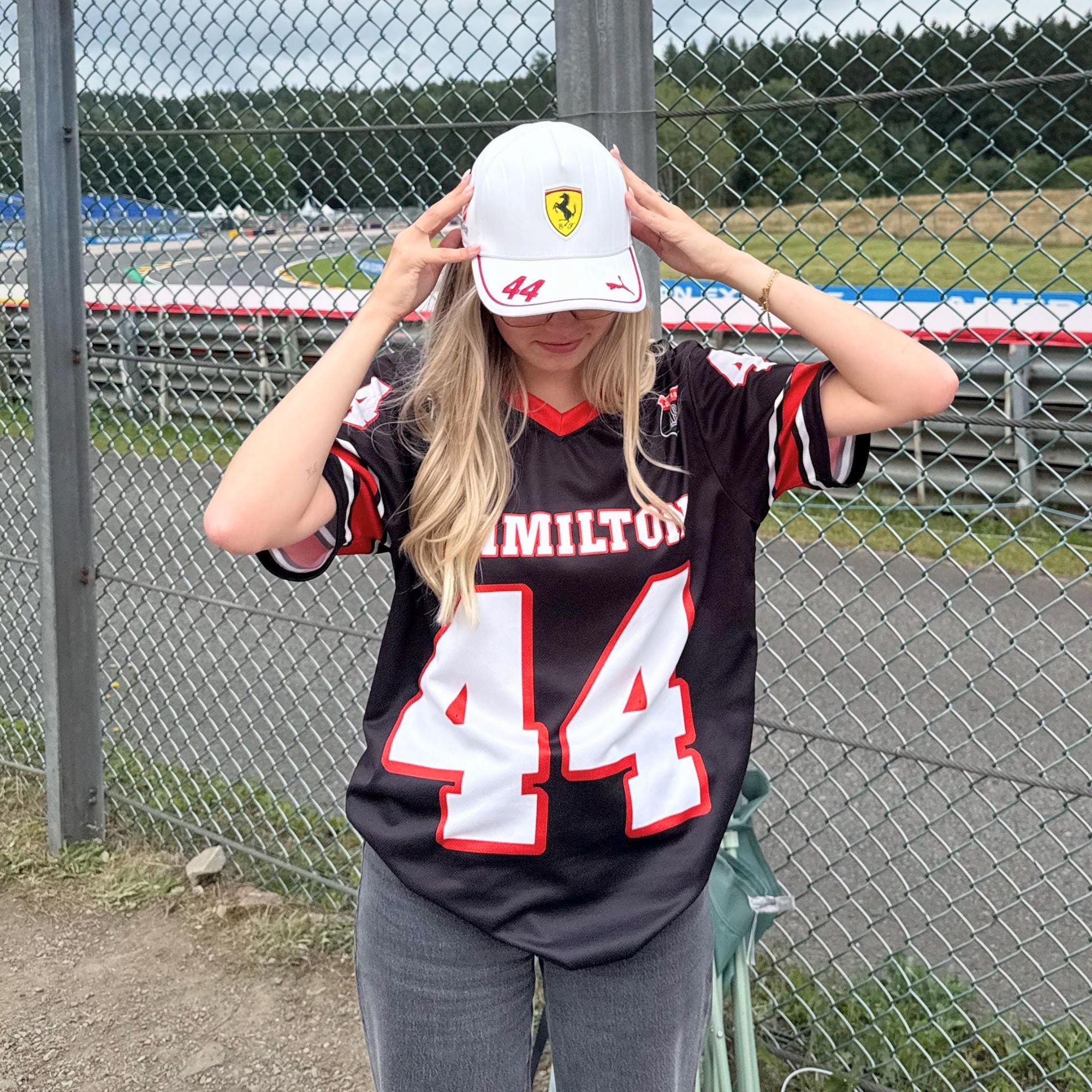 Person wearing a black sports jersey with number 44 and a white cap with a logo, standing in front of a chain-link fence.