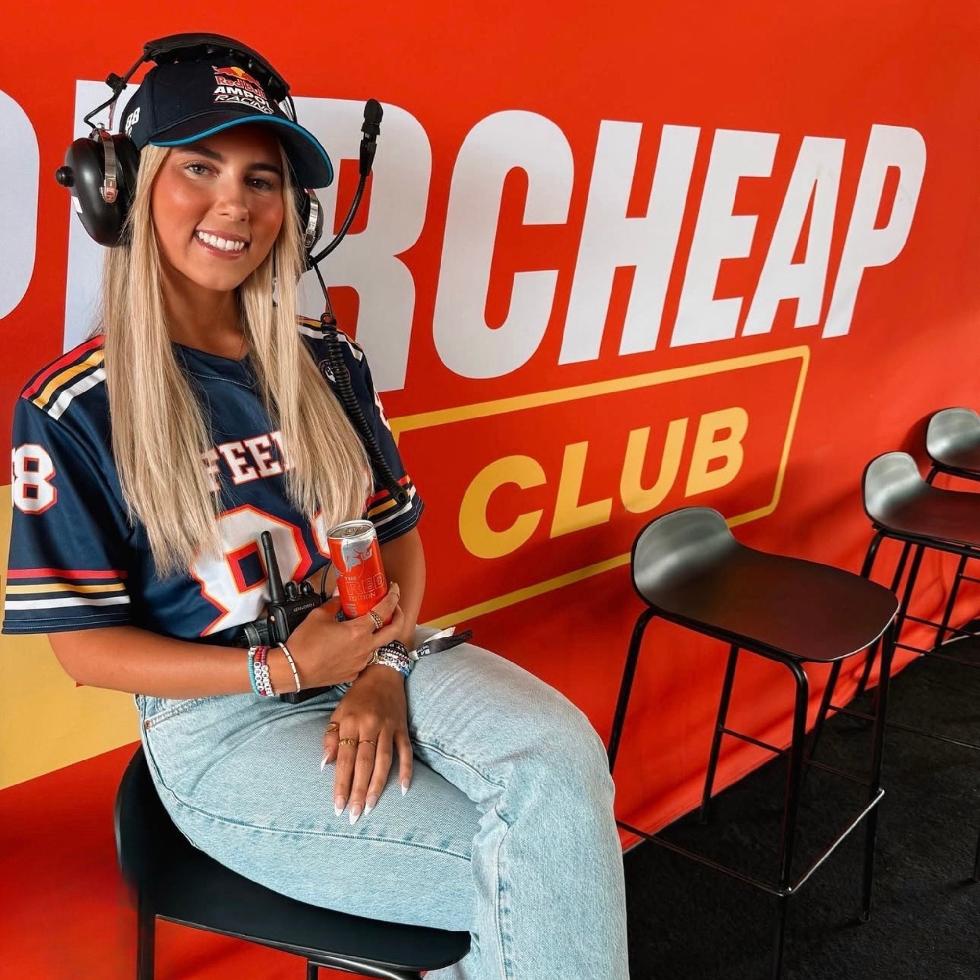 Woman sitting in a chair with a microphone, wearing headphones, in front of a red background with 'Supercheap Auto' branding.