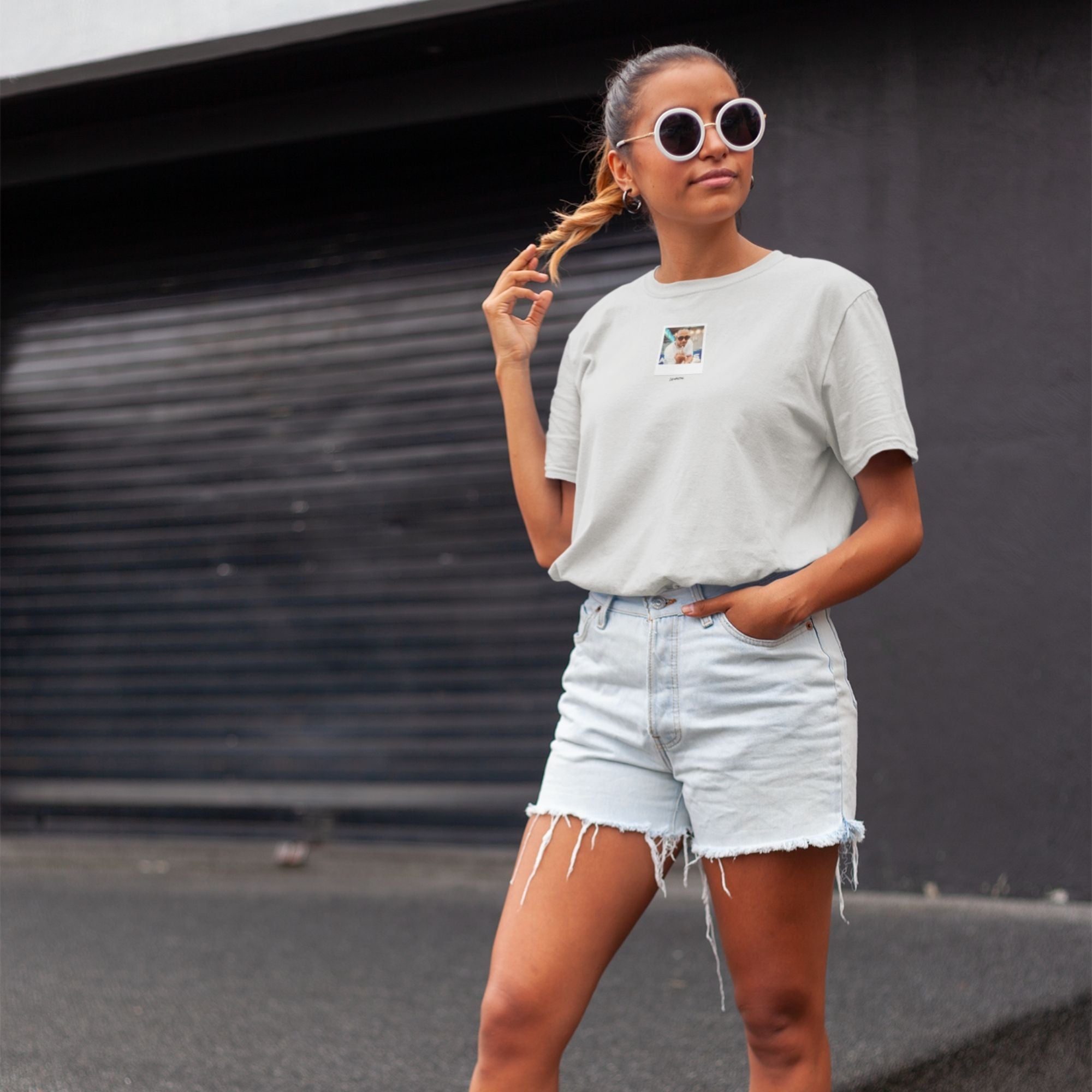 Woman wearing a light gray t-shirt and denim shorts with sunglasses, standing against a dark background.