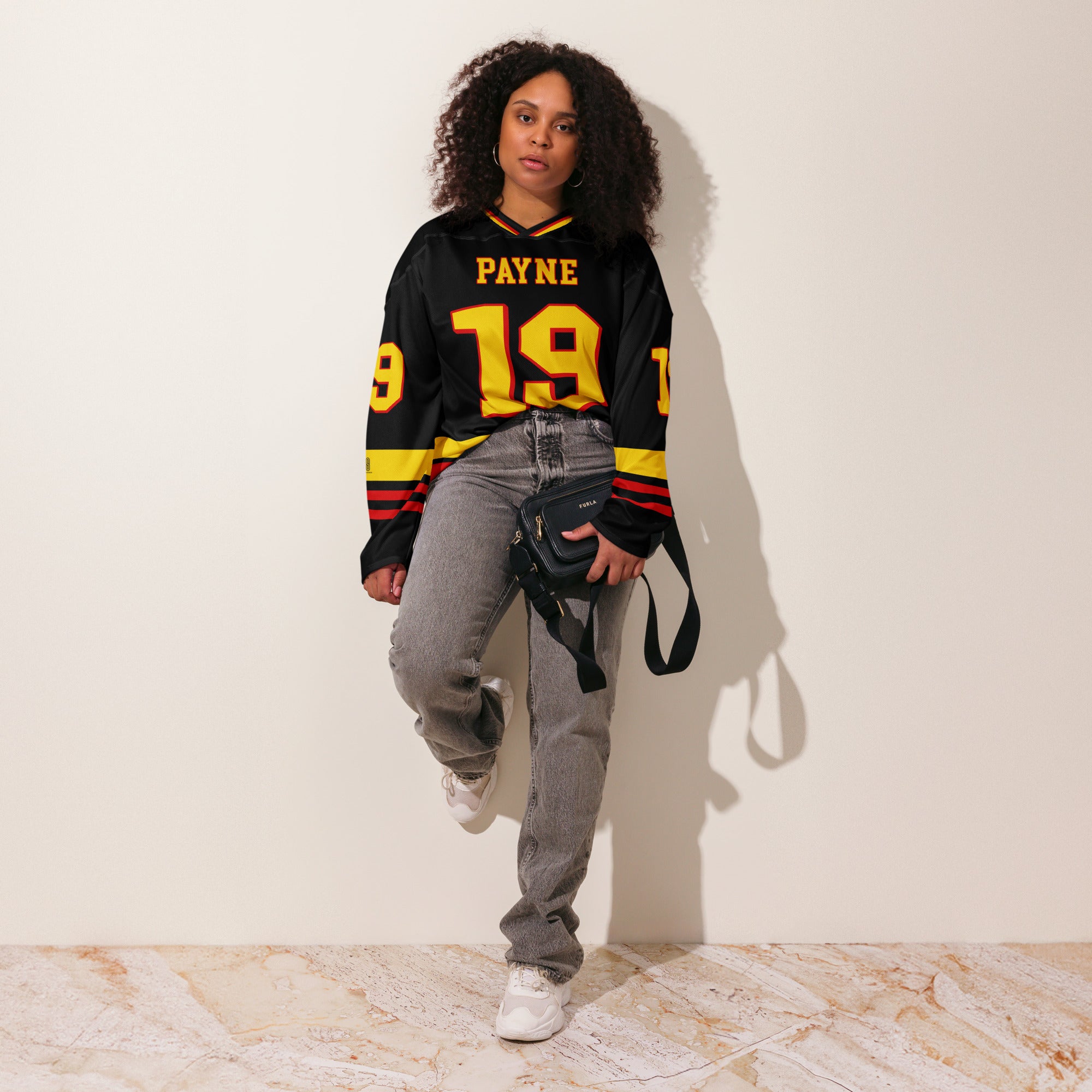 Person wearing a black sports jersey with yellow and red details, standing against a white wall.