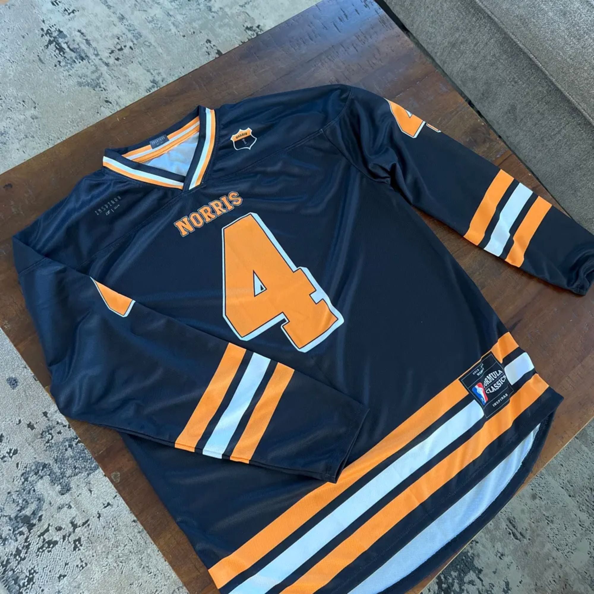 A black hockey jersey with papaya orange stripes. It has Lando Norris McLaren F1 Racing number 4 on the front and back in orange with his name on each side as well.