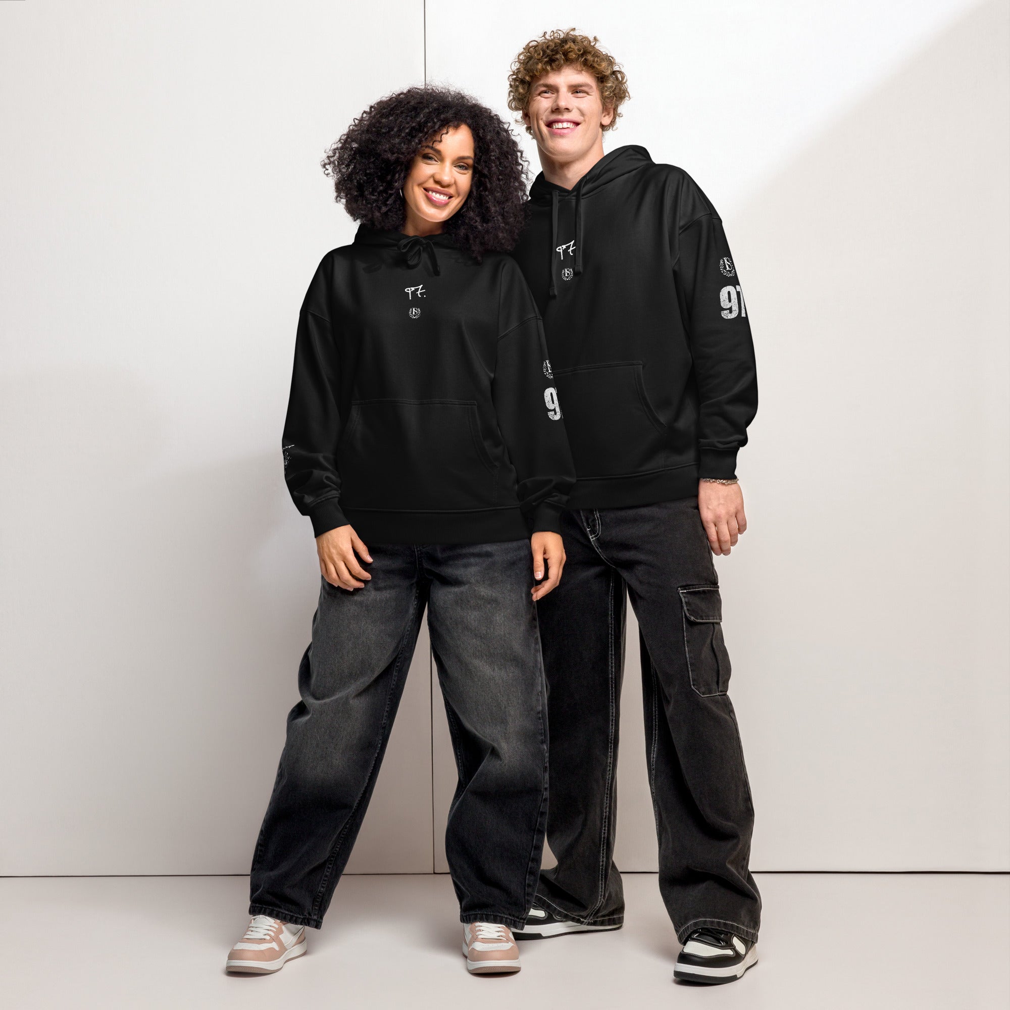 Two people wearing black hoodies and jeans standing against a white background