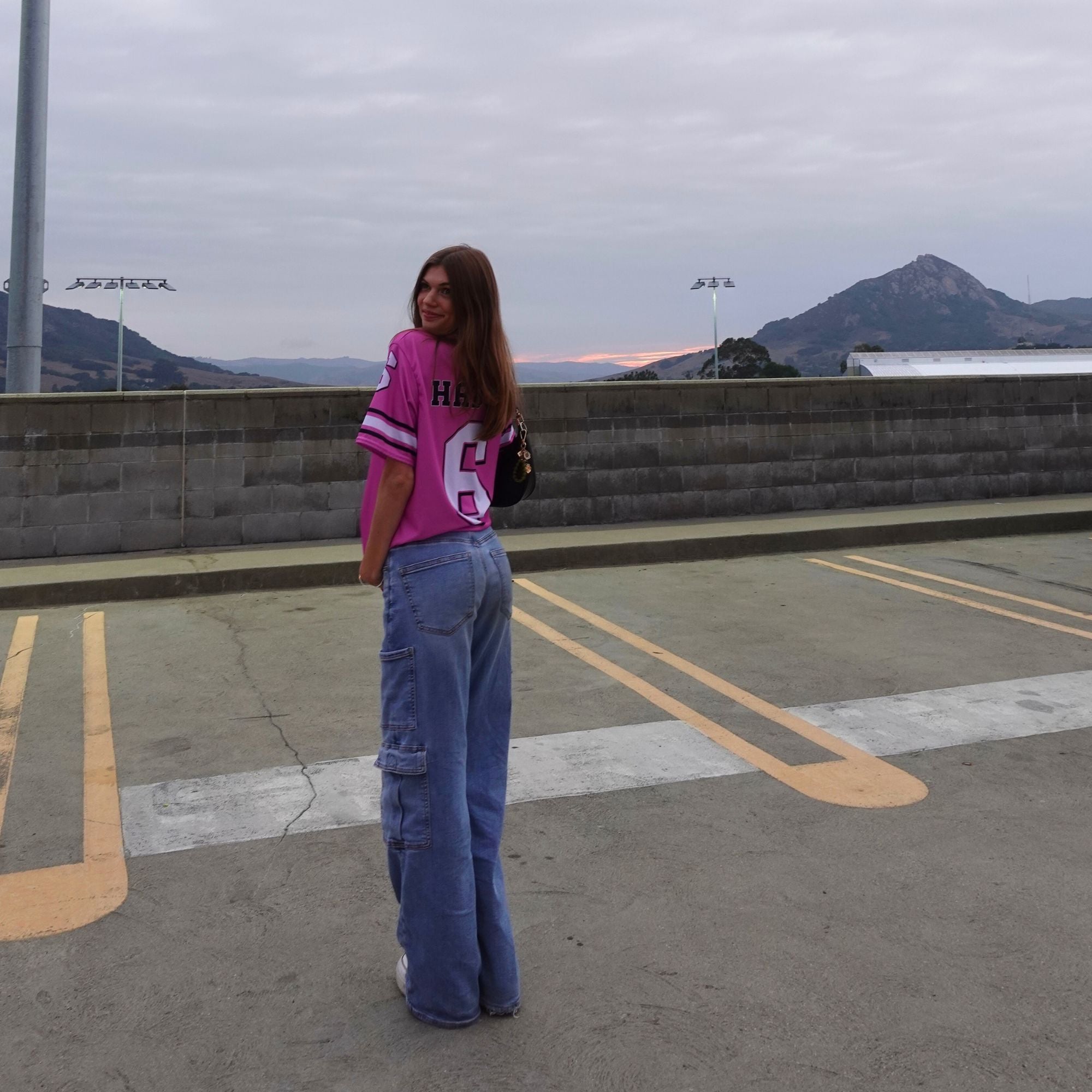 Person wearing a pink jersey with a number and blue jeans standing on a rooftop with mountains in the background.