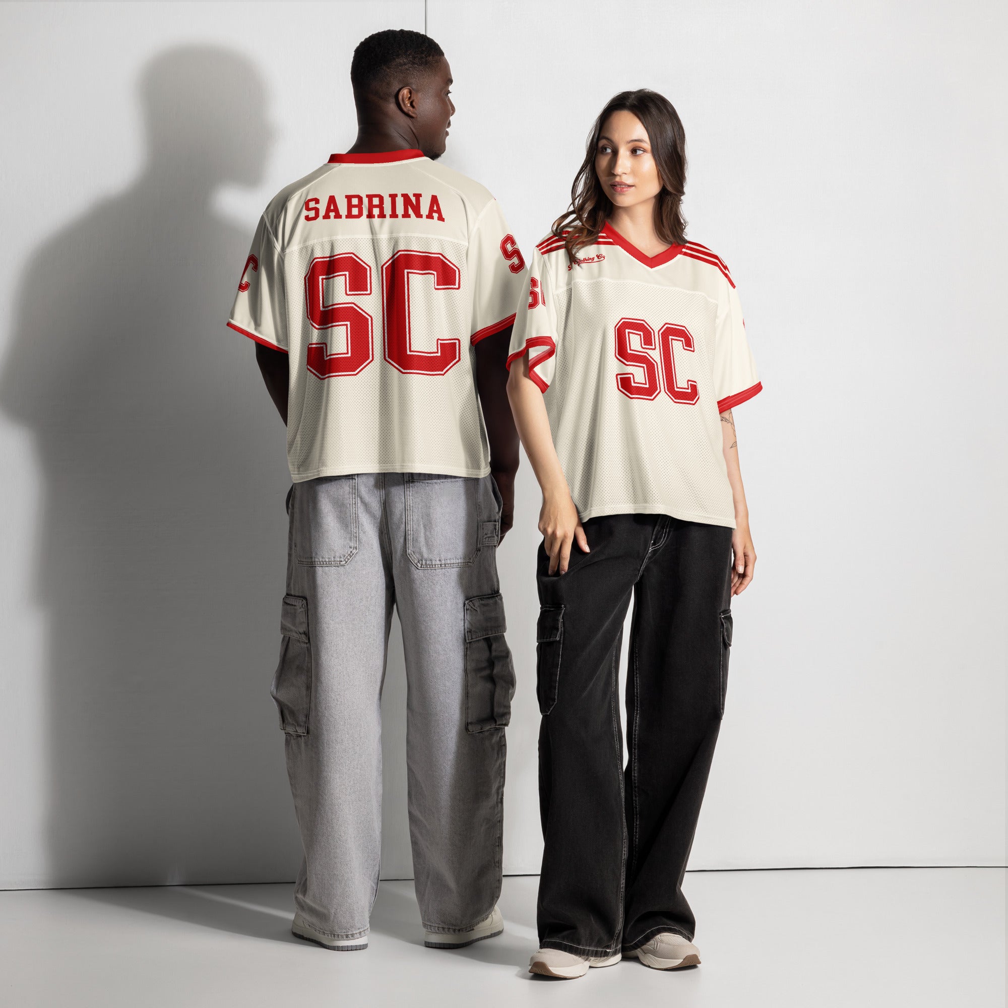 Two people wearing matching cream jerseys with red text and 'SC' logo on a plain background